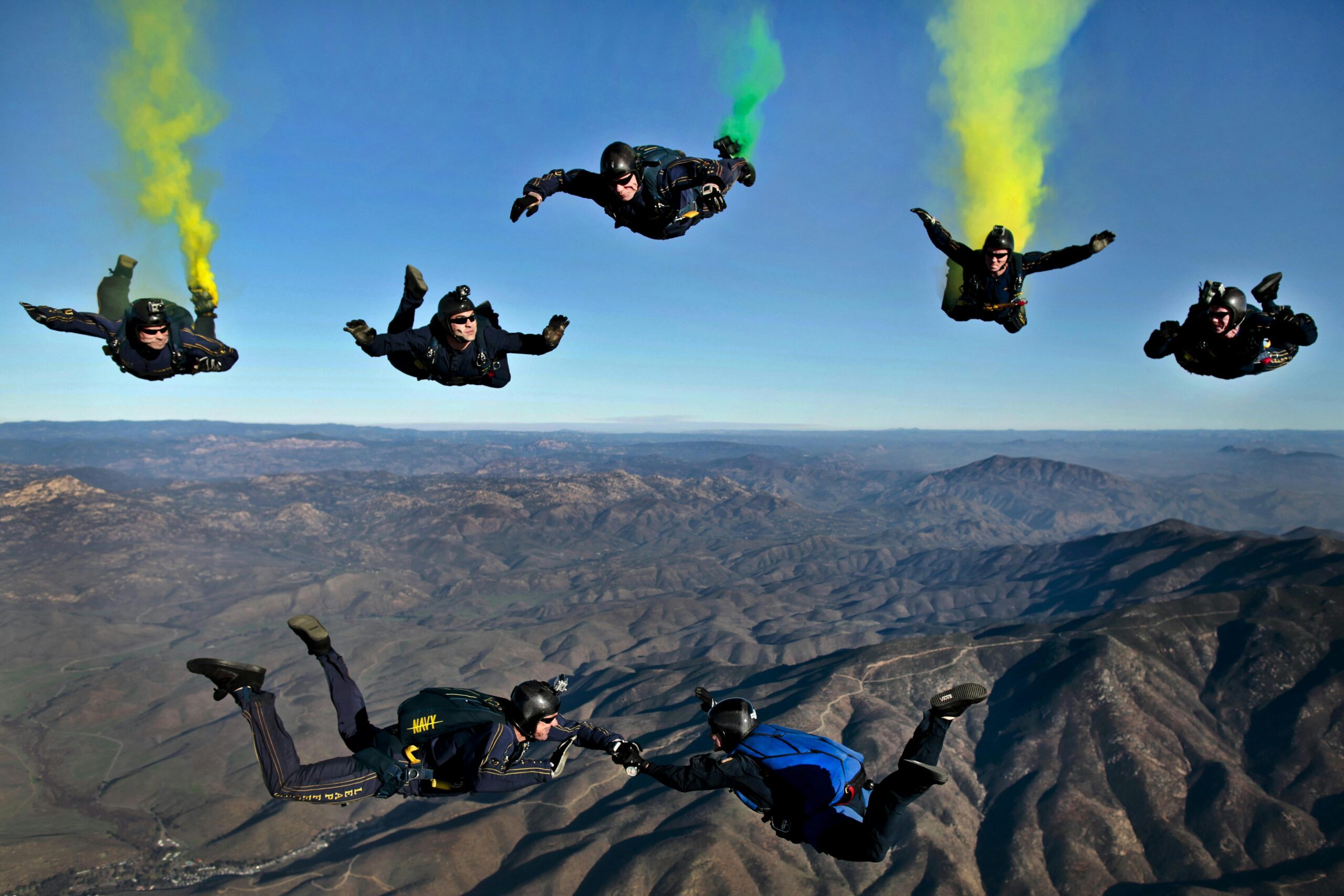 Dynamic aerial shot of skydivers performing stunts with colorful smoke trails over mountainous terrain.