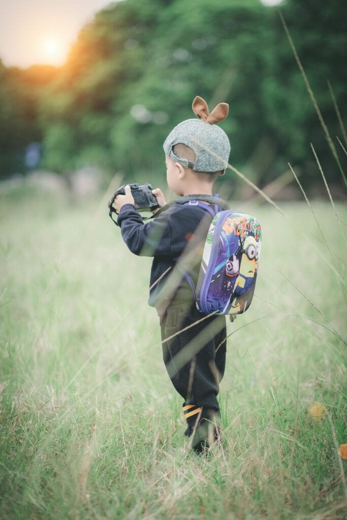 Child with backpack and camera captures nature in grassy field during sunset.