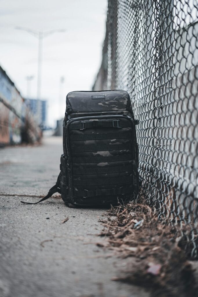 A rugged black backpack leaning against a chain link fence on an urban sidewalk.