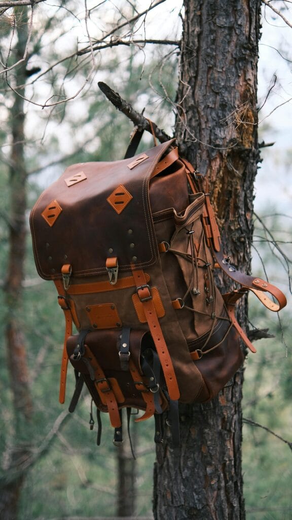A rustic leather backpack hangs on a tree in a forest setting, ideal for outdoor adventure themes.