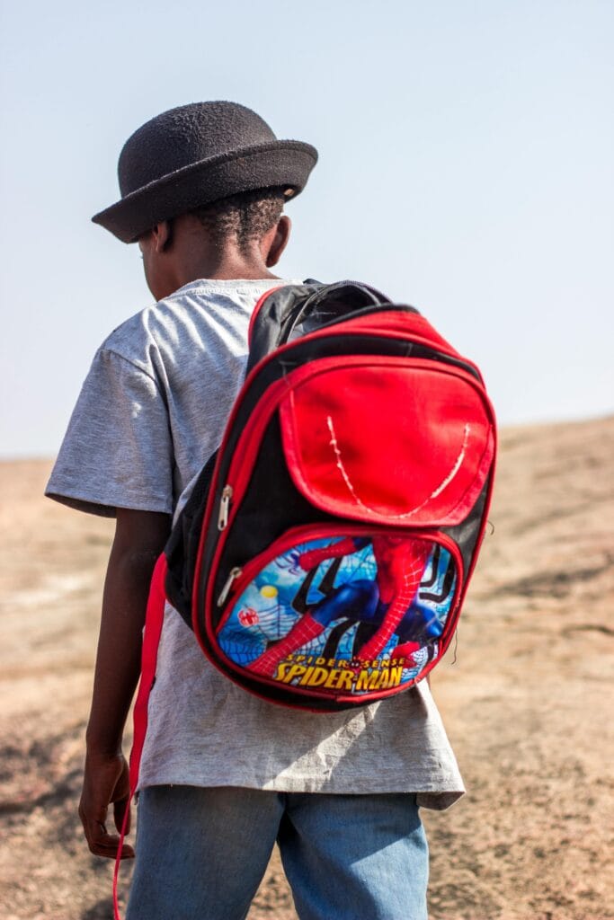 A young boy wearing a spiderman backpack stands outdoors, seen from behind.