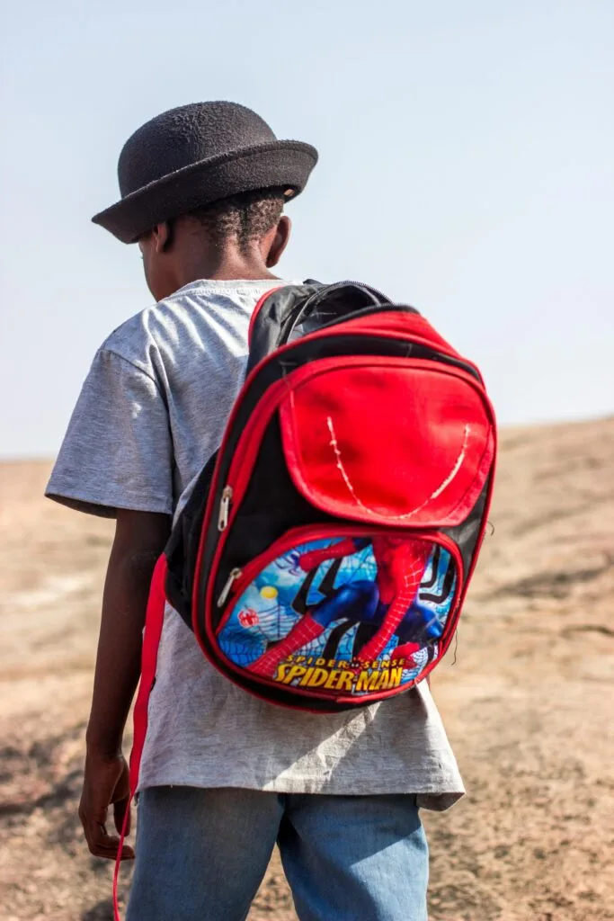 A young boy wearing a spiderman backpack stands outdoors, seen from behind.