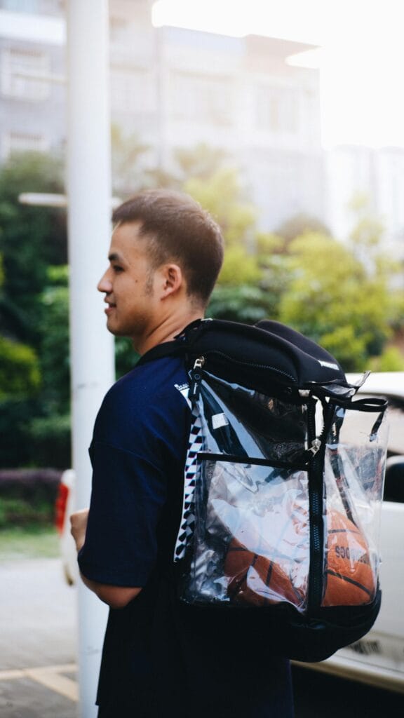 Smiling man carrying a clear backpack with a basketball on a sunny day.