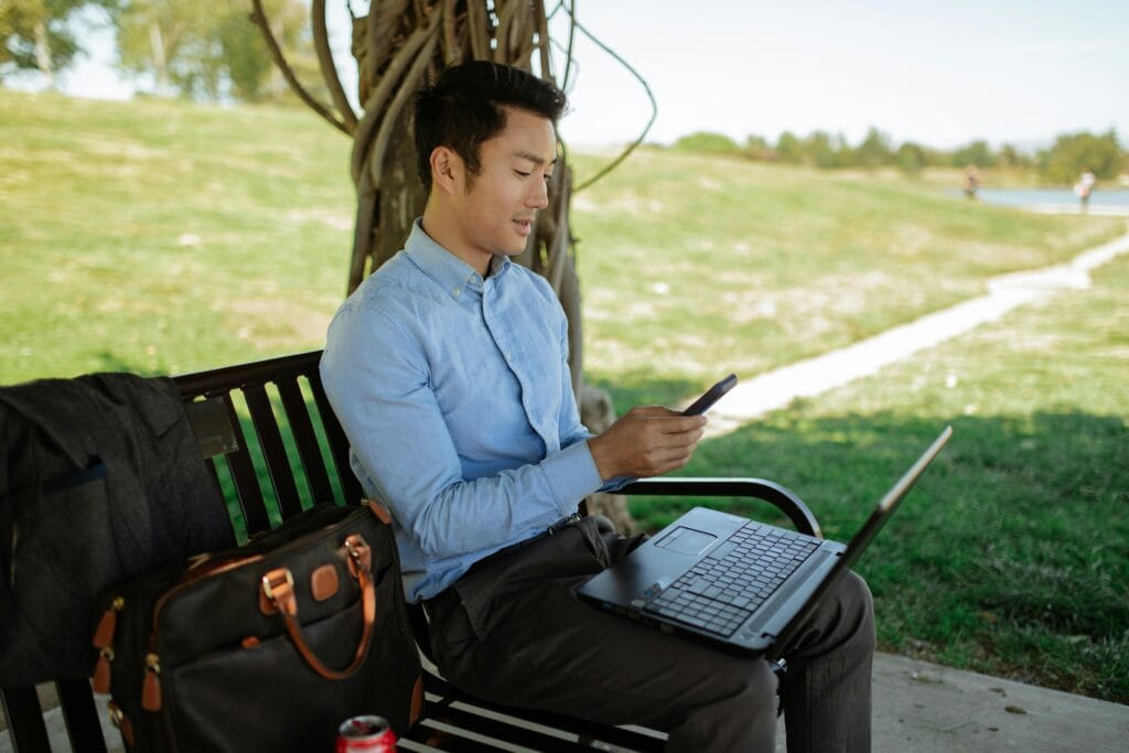 Asian man working on a laptop outdoors while checking his cellphone. Remote work lifestyle.