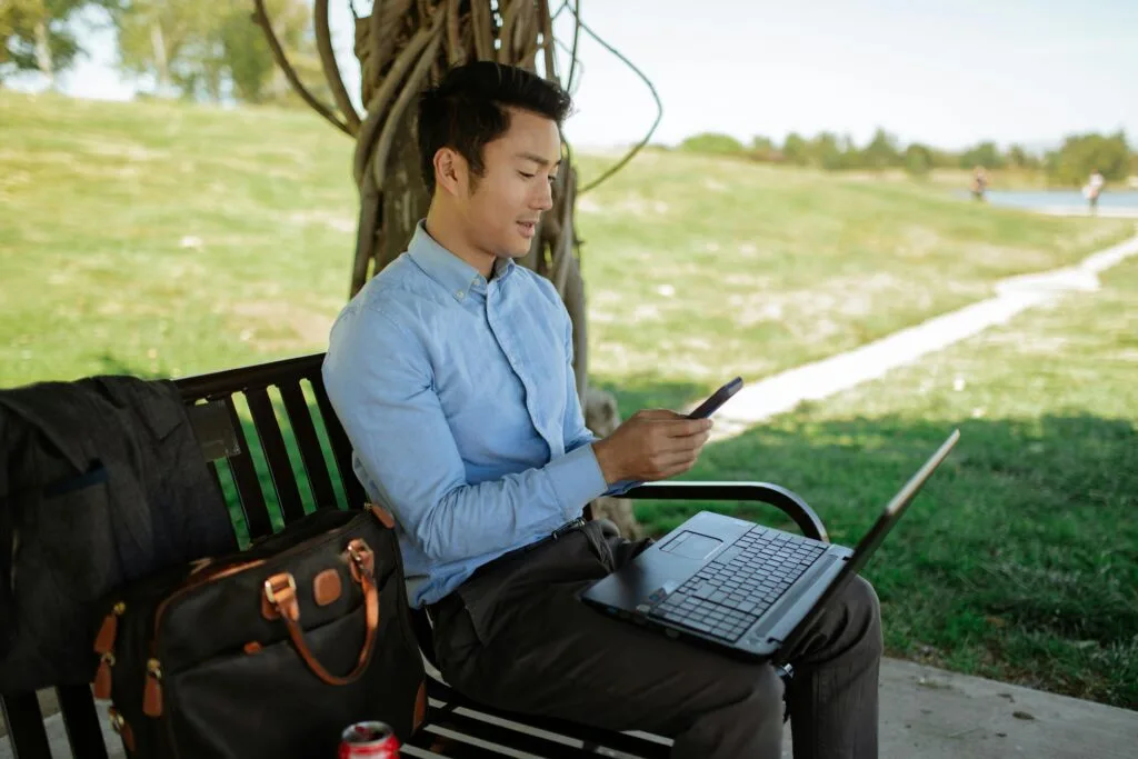 Asian man working on a laptop outdoors while checking his cellphone. Remote work lifestyle.
